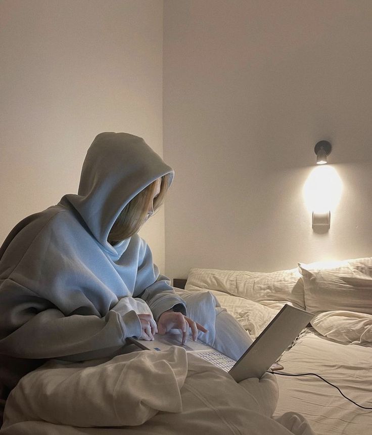 a person sitting on a bed with a laptop in front of them while wearing a hoodie