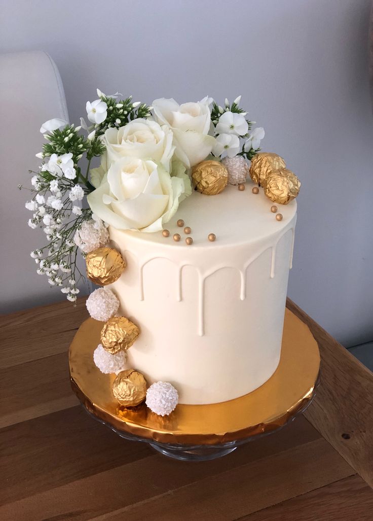 a white cake with gold decorations and flowers on the top is sitting on a wooden table