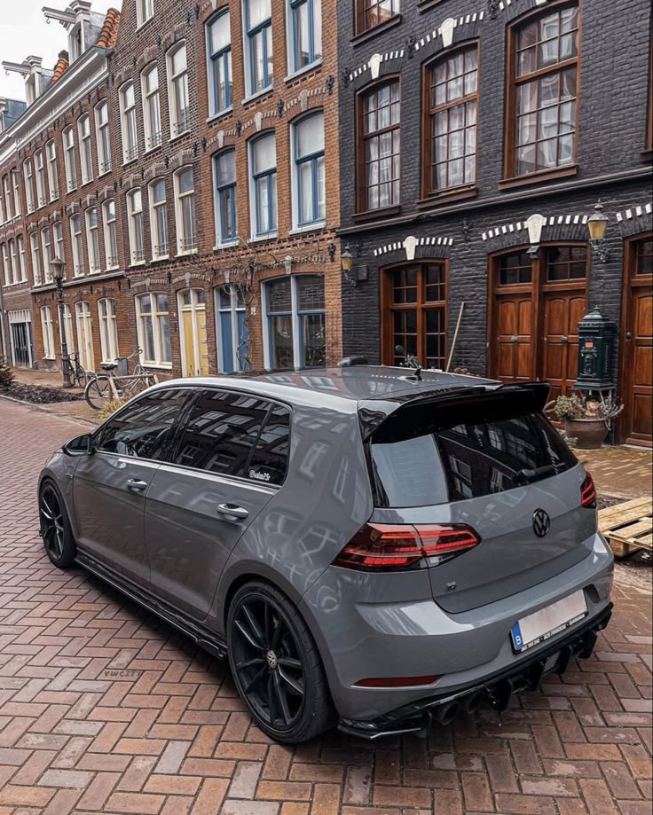 a grey car parked on the side of a brick road next to tall brown buildings