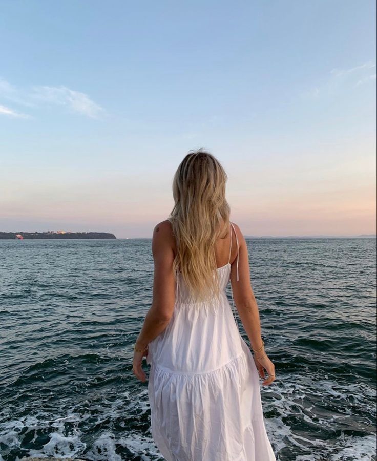 a woman in a white dress standing on the edge of a boat looking out at the water