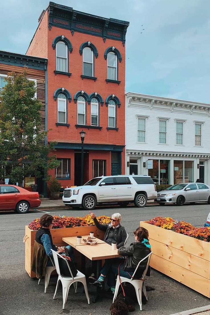 three people sitting at a table in front of some flowers on the side of the road