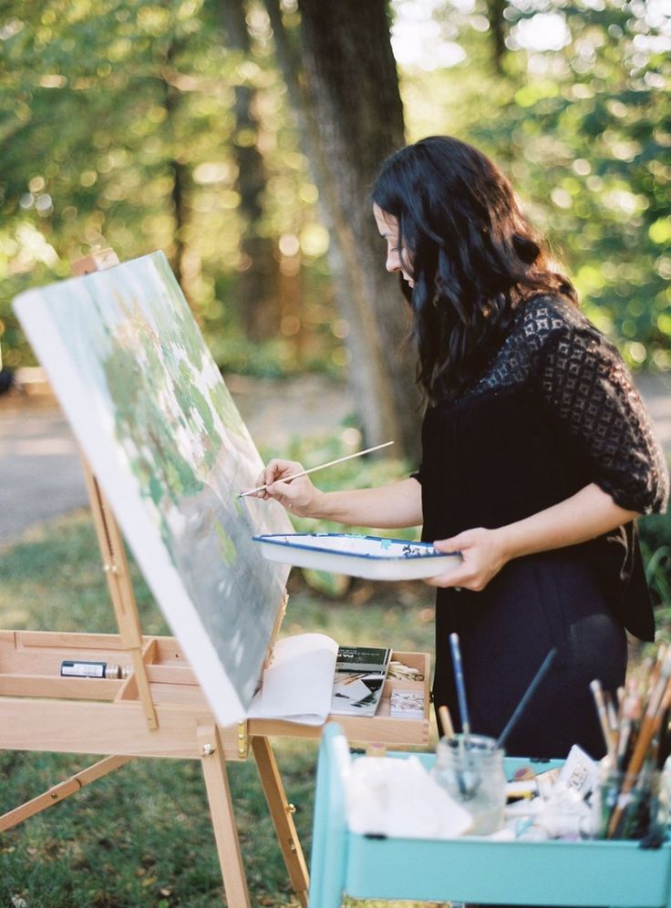 a woman is painting on an easel in the woods with her hands holding a paintbrush