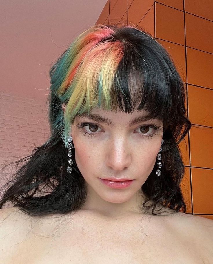 a woman with multi - colored hair is posing in front of an orange brick wall