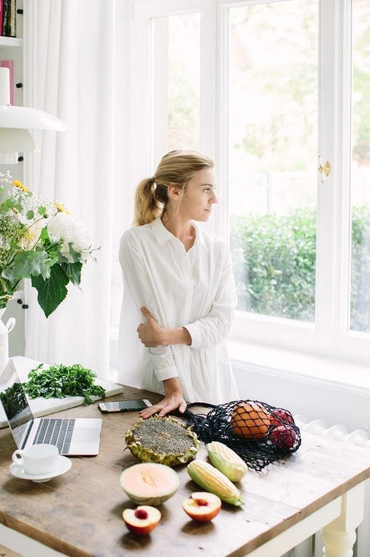 a woman standing in front of a window with fruit on the table next to her