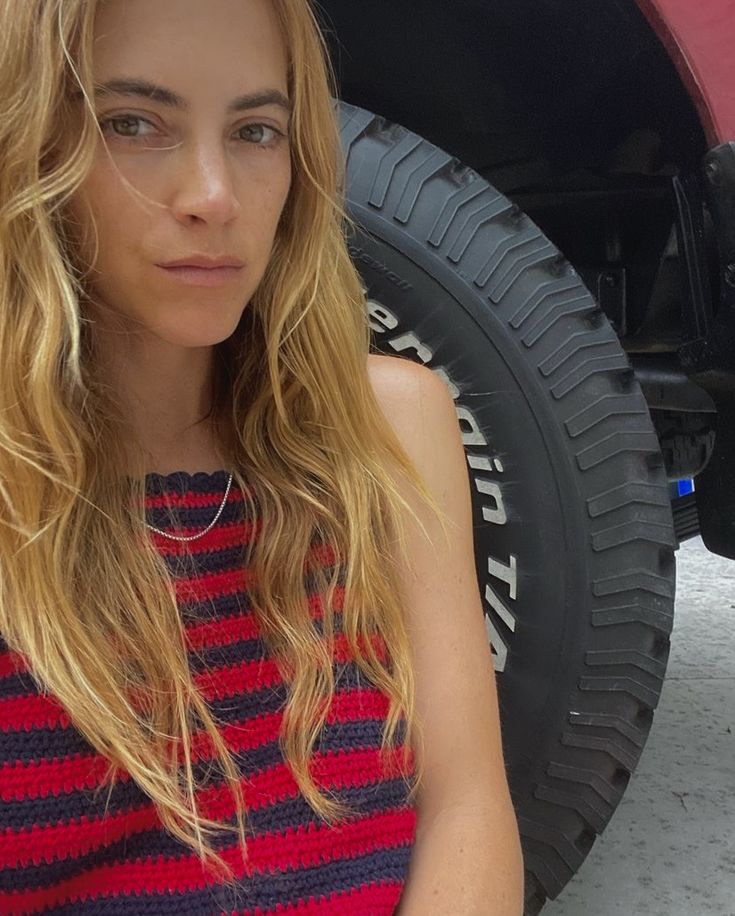 a woman sitting in front of a red car with her hand on her hip and looking at the camera