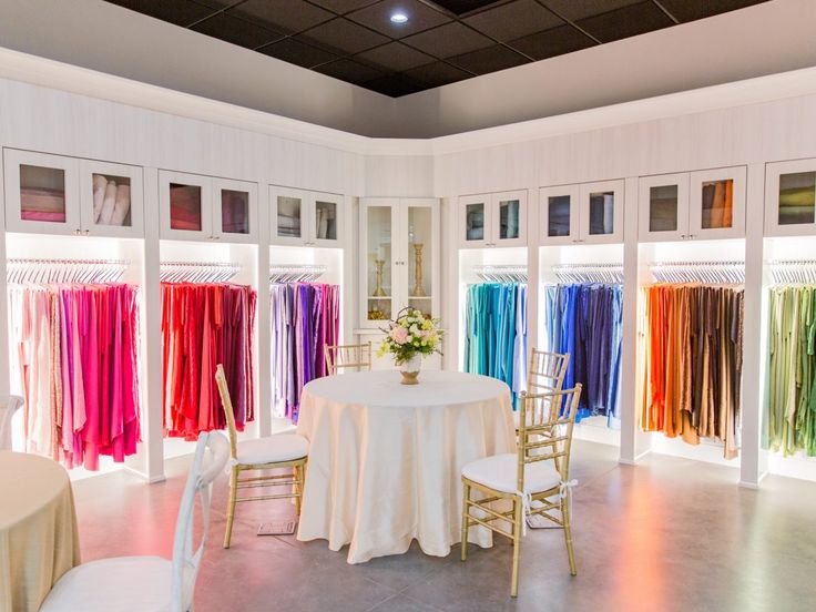 a room filled with tables and chairs covered in white cloths next to colorful dresses