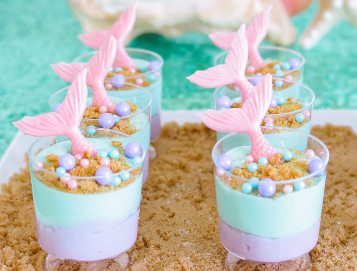 three little mermaid desserts in small glasses on a tray with sand and sea shells