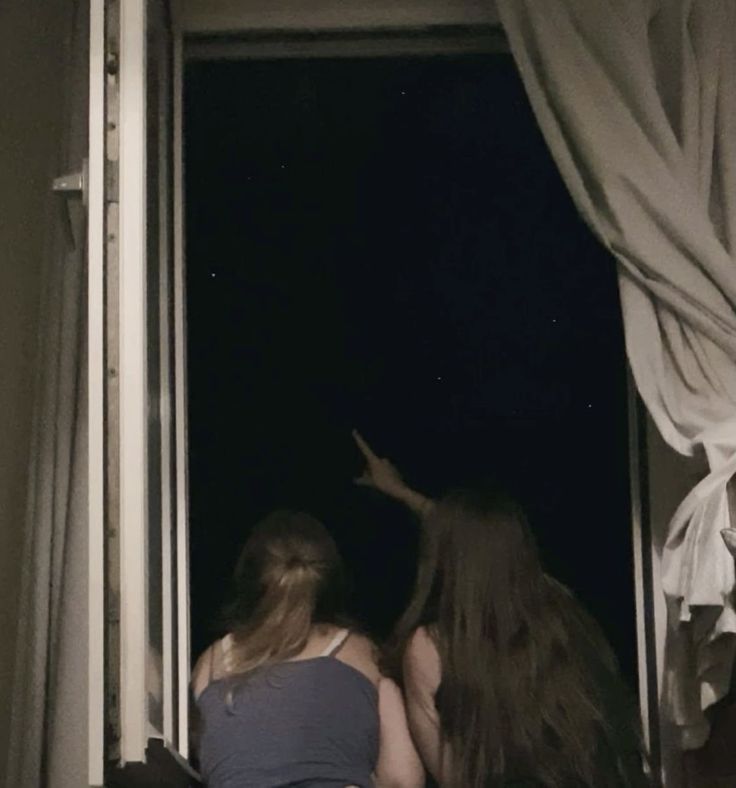 two girls are looking out the window at the dark night sky and one girl is reaching for something