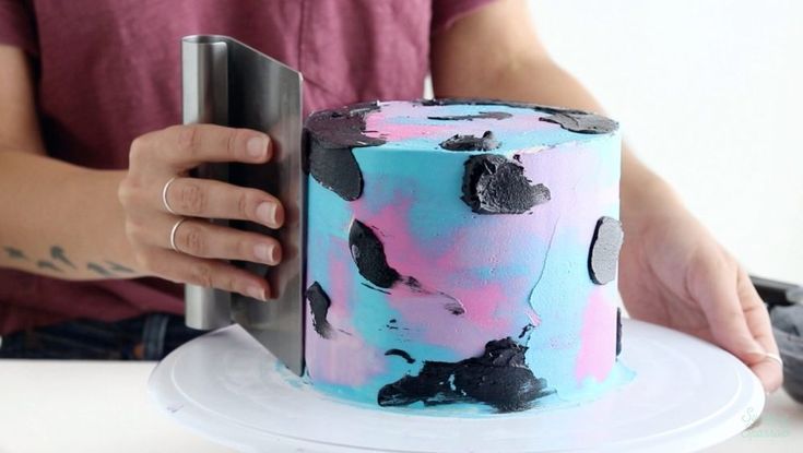 a person is holding a knife over a colorful cake on a white plate with pink, blue and black icing