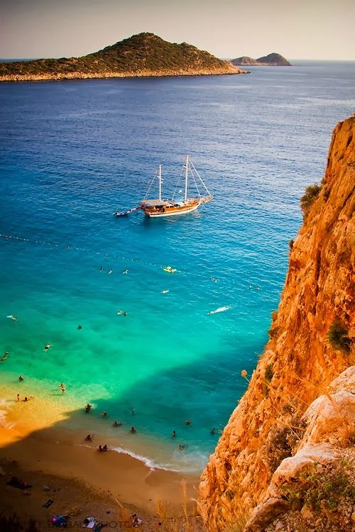 people are swimming in the blue water near a rocky cliff with a boat on it