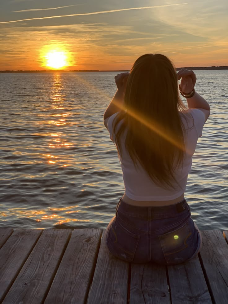 a woman sitting on a dock looking out at the water with the sun setting behind her