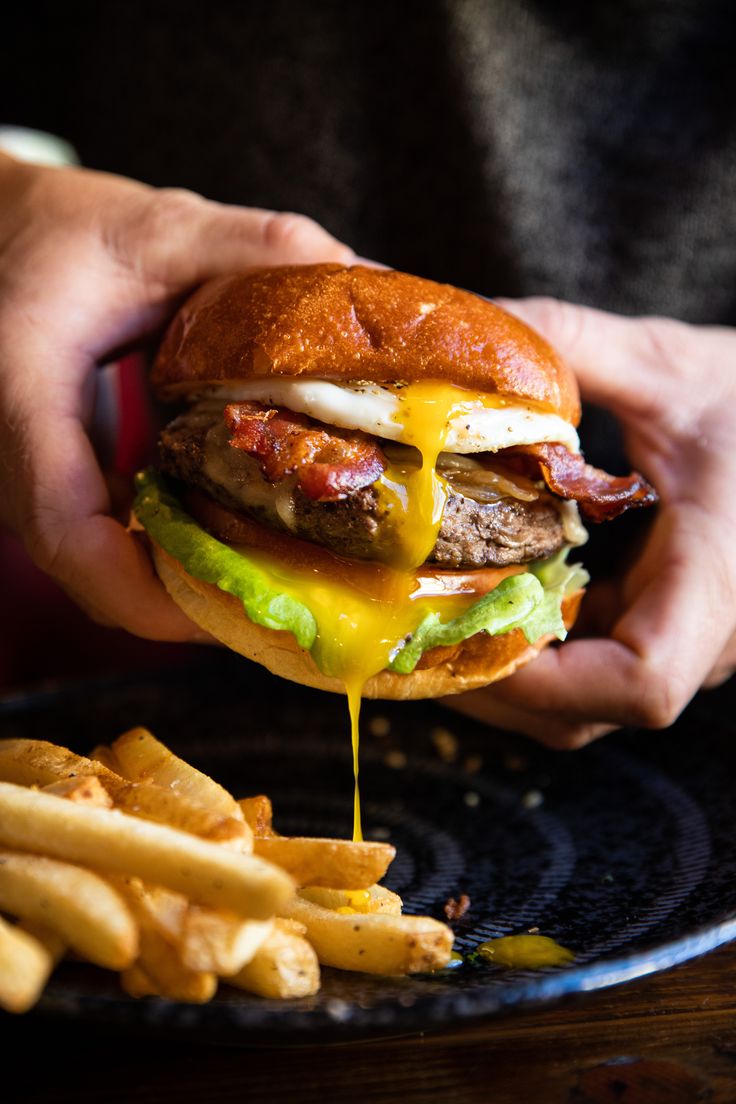 a person holding a hamburger with cheese and lettuce next to french fries on a plate