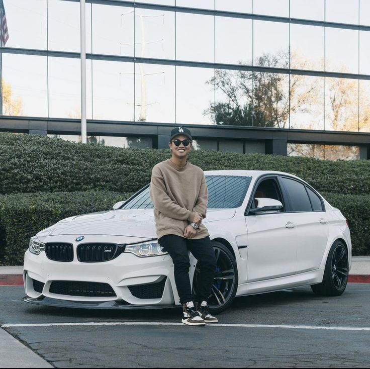 a man sitting on the hood of a white car in front of a tall building