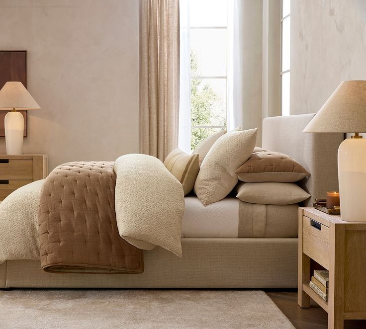 a bed with pillows and blankets on top of it next to a night stand in front of a window