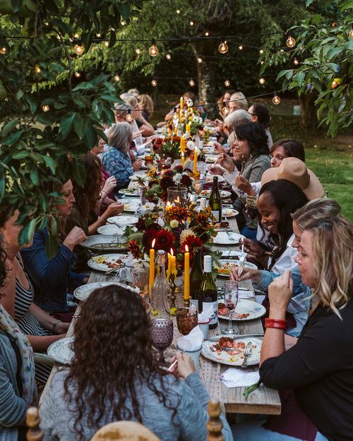 a group of people sitting around a long table with food and drinks on it, surrounded by greenery