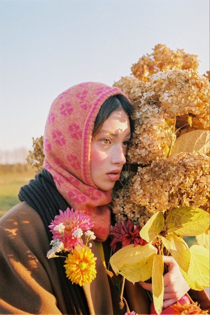 a woman wearing a pink head scarf holding flowers in front of her face and looking off into the distance