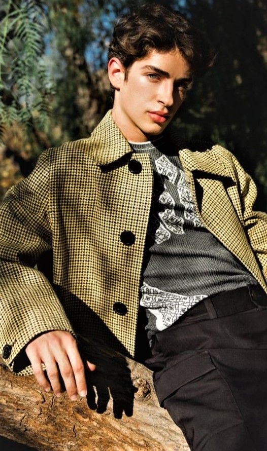 a young man sitting on top of a tree branch wearing a jacket and tie with his hands in his pockets