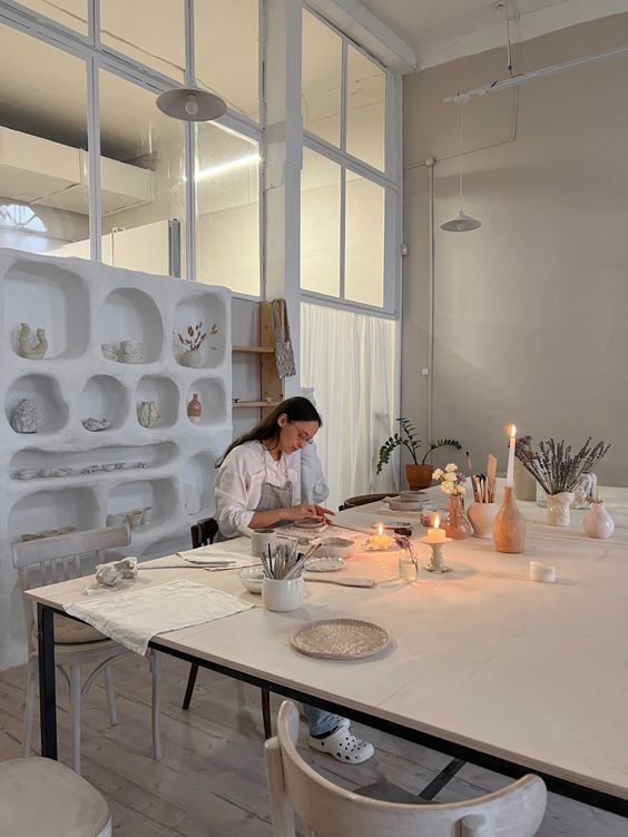 a woman sitting at a table in front of a white wall with shelves on it