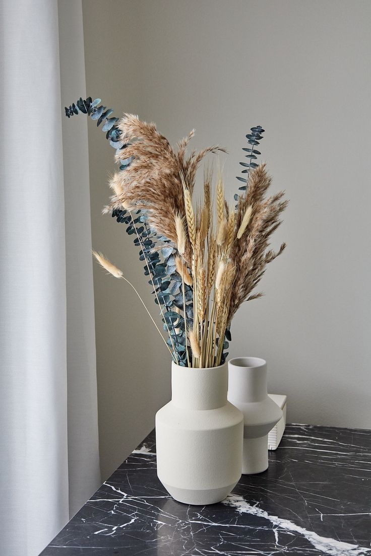 two vases with dried plants in them on a marble countertop next to a window