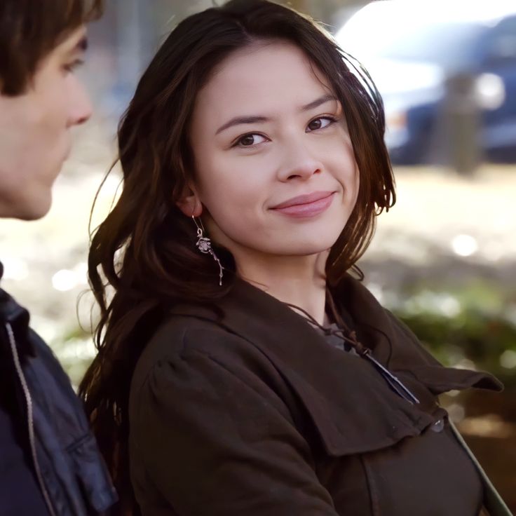 a man and woman standing next to each other in front of a tree with cars behind them