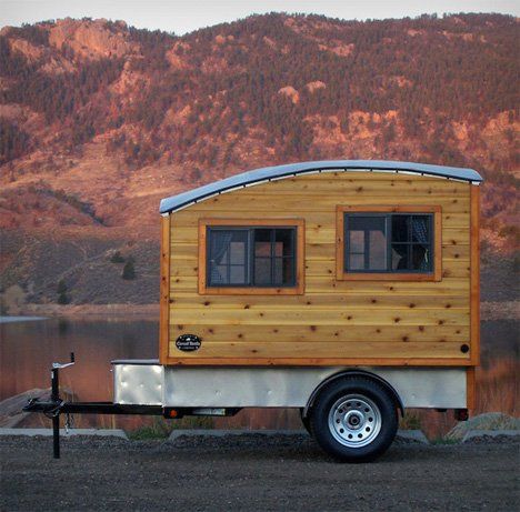 a tiny house on wheels parked next to a lake with mountains in the back ground