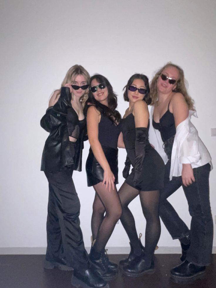 four women posing for the camera in front of a white wall wearing black clothing and sunglasses