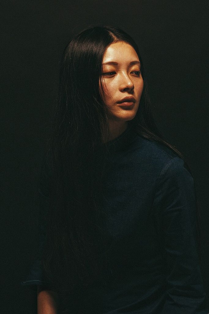 a woman with long black hair standing in front of a dark background and looking off to the side