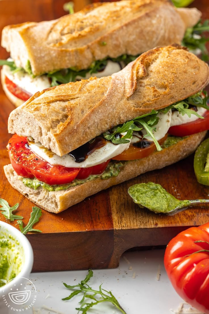 two sub sandwiches sitting on top of a wooden cutting board next to tomatoes and guacamole