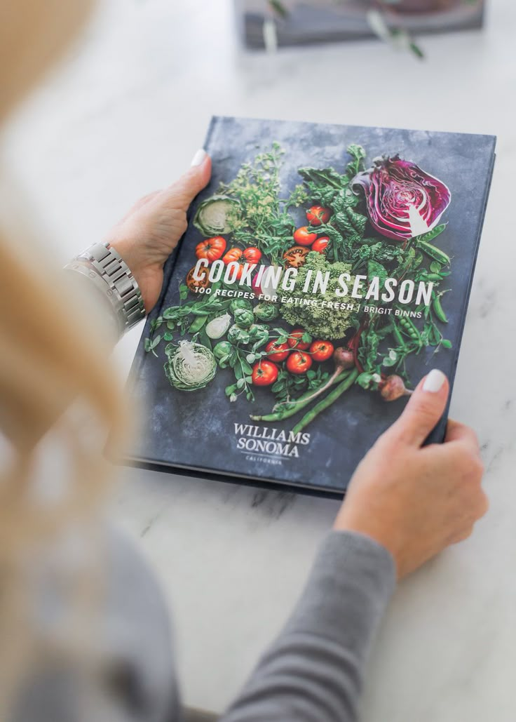 the cookbook is being held up by a woman's hands while she sits at a table