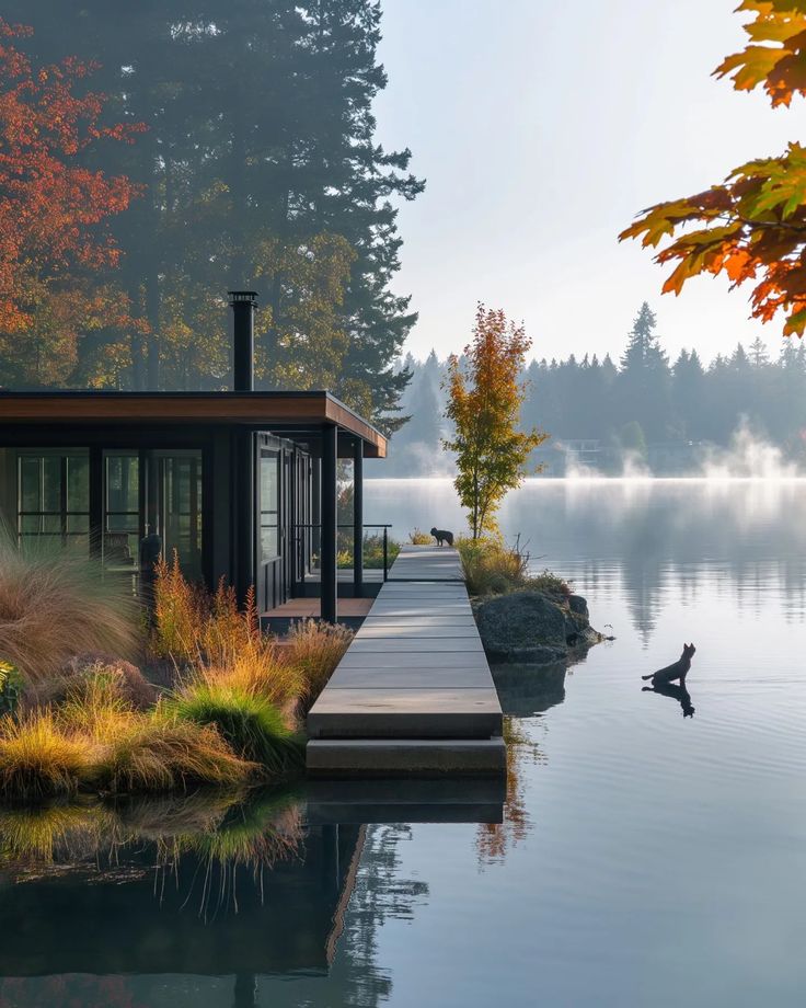 a house on the edge of a body of water with trees in the background and a dock leading to it