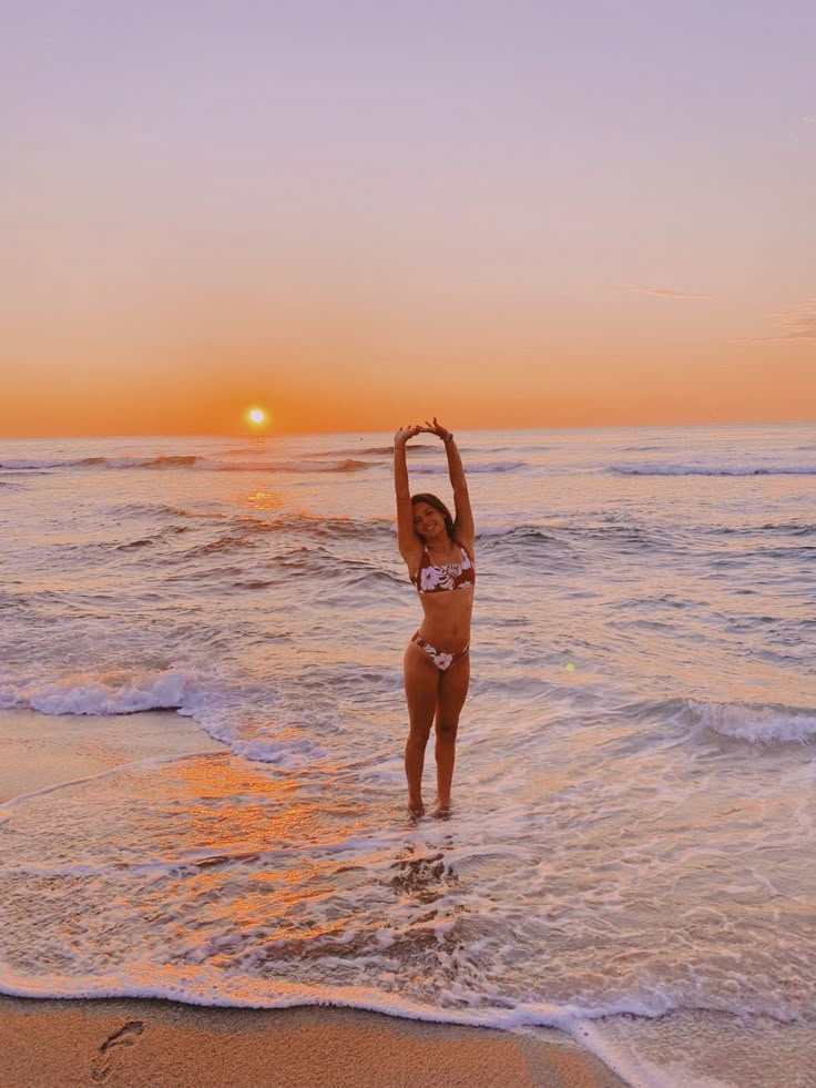 a woman standing in the ocean at sunset with her arms up and hands above her head