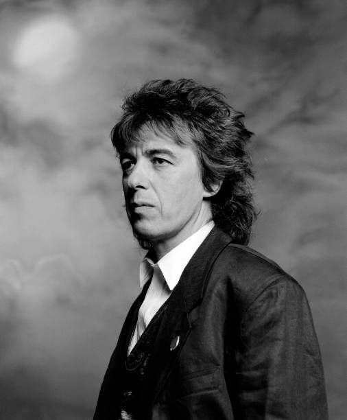 a black and white photo of a man with long hair wearing a suit, standing in front of a cloudy sky