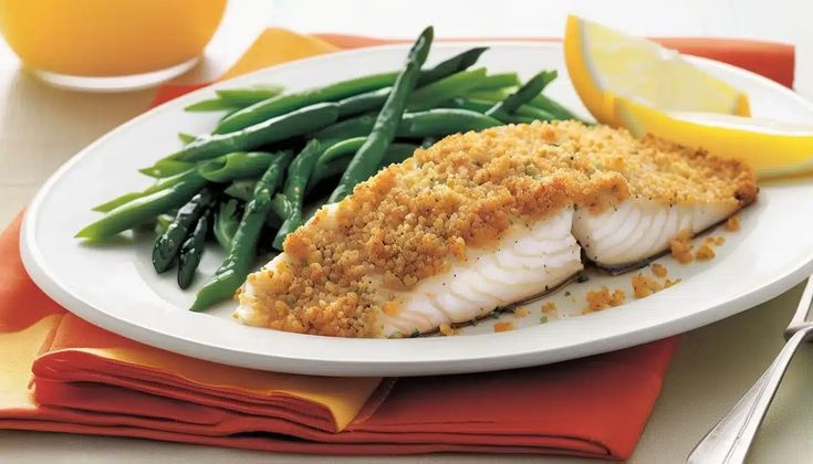 a white plate topped with fish and green beans next to lemon wedges on a table