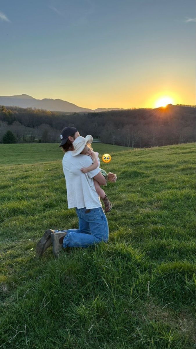 a woman holding a baby in her arms while standing on top of a lush green field