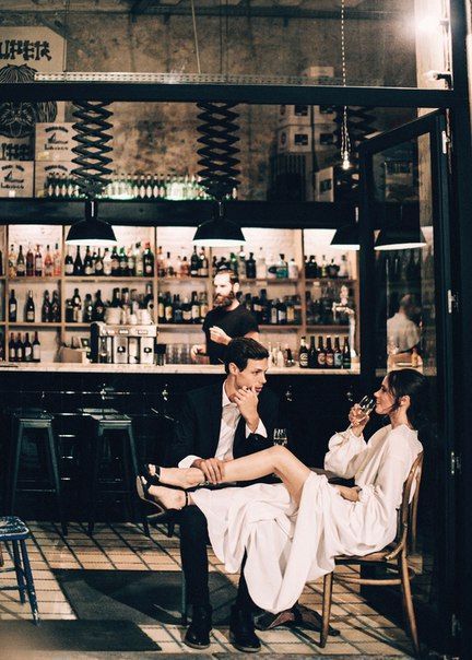 two men sitting in chairs at a bar with bottles on the wall and people standing around