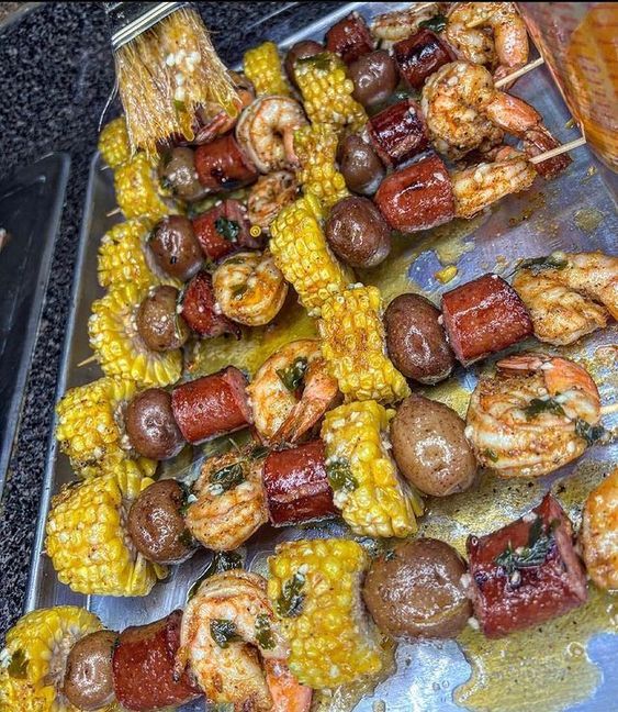 several skewered meats and vegetables are on a metal tray next to corn