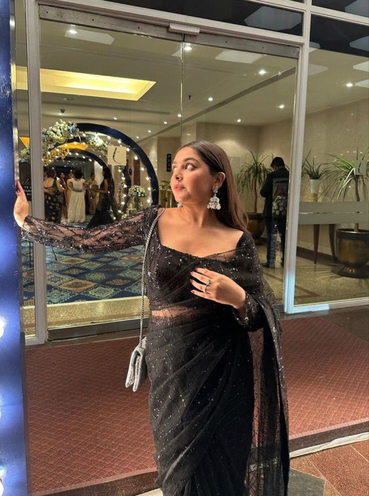 a woman standing in front of a store window wearing a black sari and holding her hand out