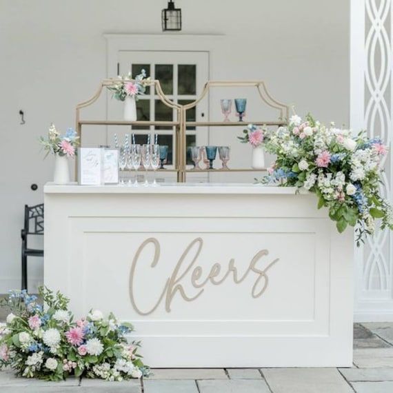 an outdoor bar with flowers and greenery on the counter at a wedding reception in front of a white building