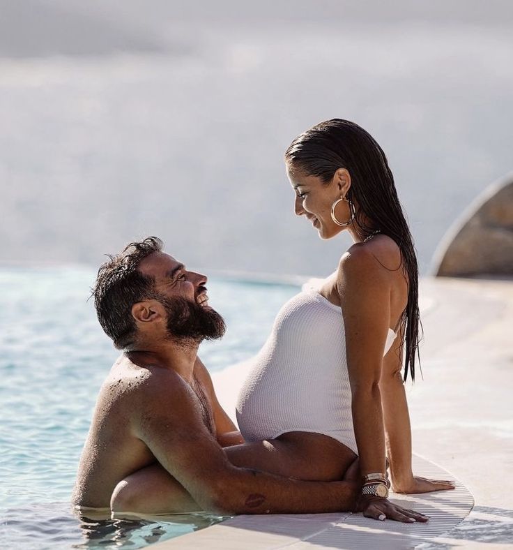 a man and woman sitting on the edge of a swimming pool looking at each other