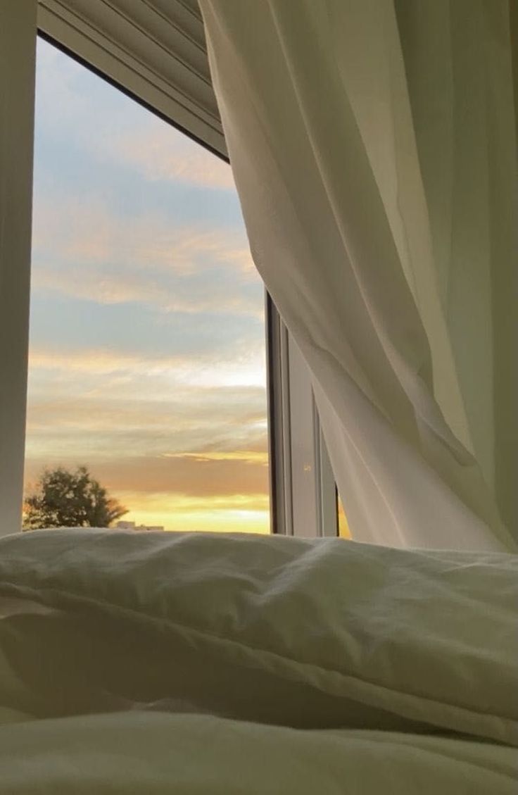 an open window with curtains in front of it and the sun setting over trees outside