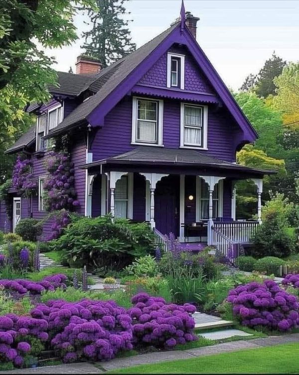 a purple house with lots of flowers in front of it and trees around the yard