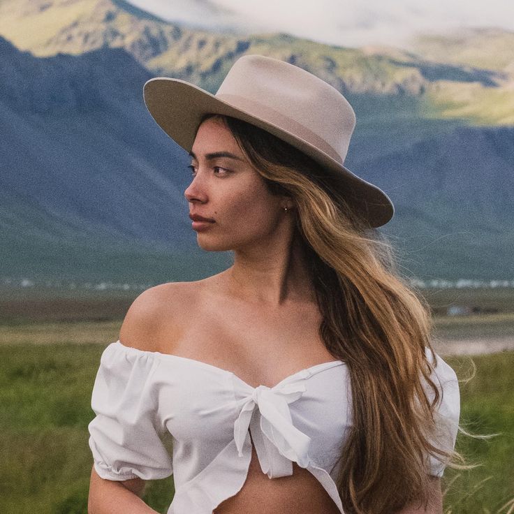 a woman with long hair wearing a white top and hat standing in front of mountains