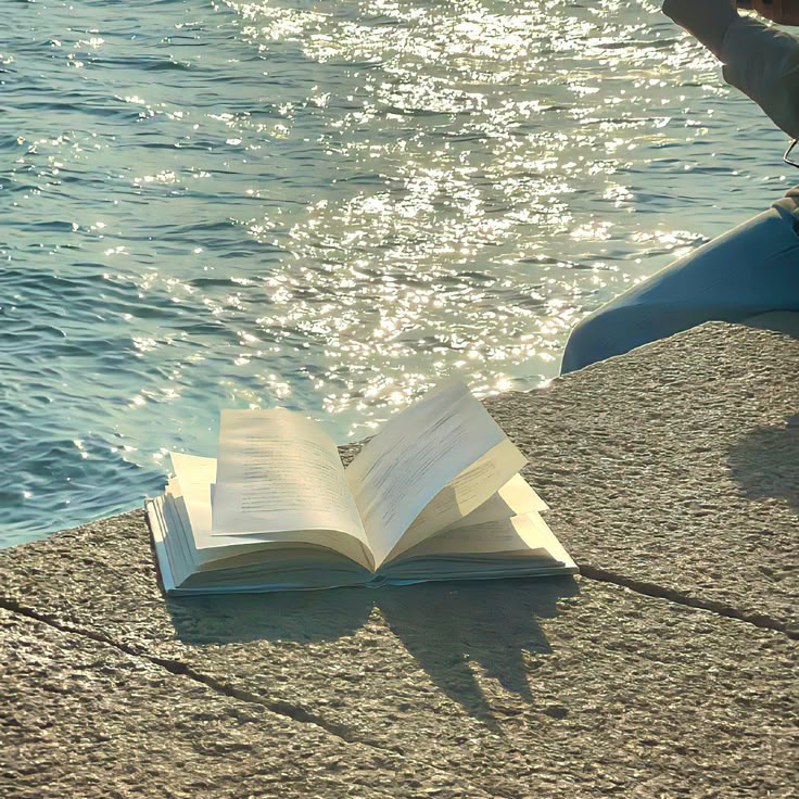 an open book sitting on the edge of a body of water next to a person