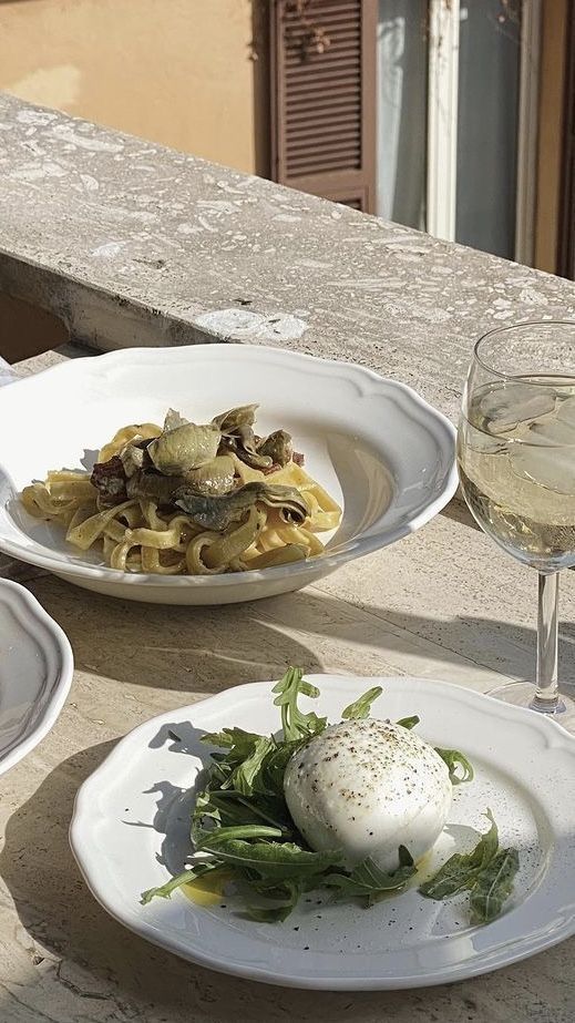 two white plates with food on them sitting on a table next to a glass of wine