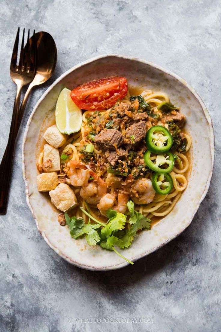 a white bowl filled with noodles and vegetables next to a fork on a gray surface