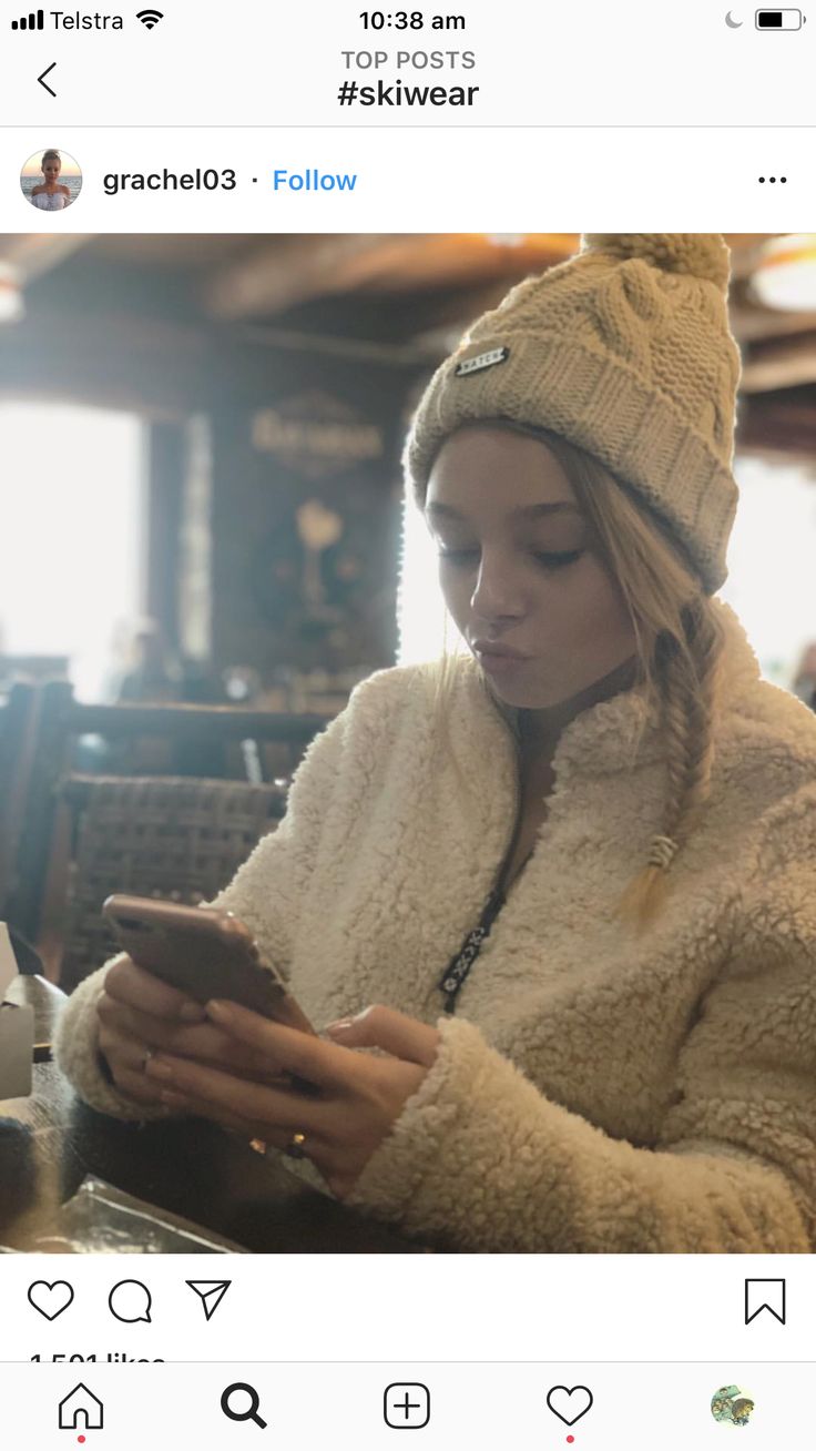 a woman sitting at a table looking at her cell phone while wearing a hat and coat