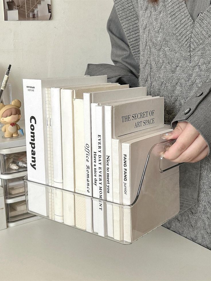 a woman holding a stack of books in front of a white desk with several other books on it