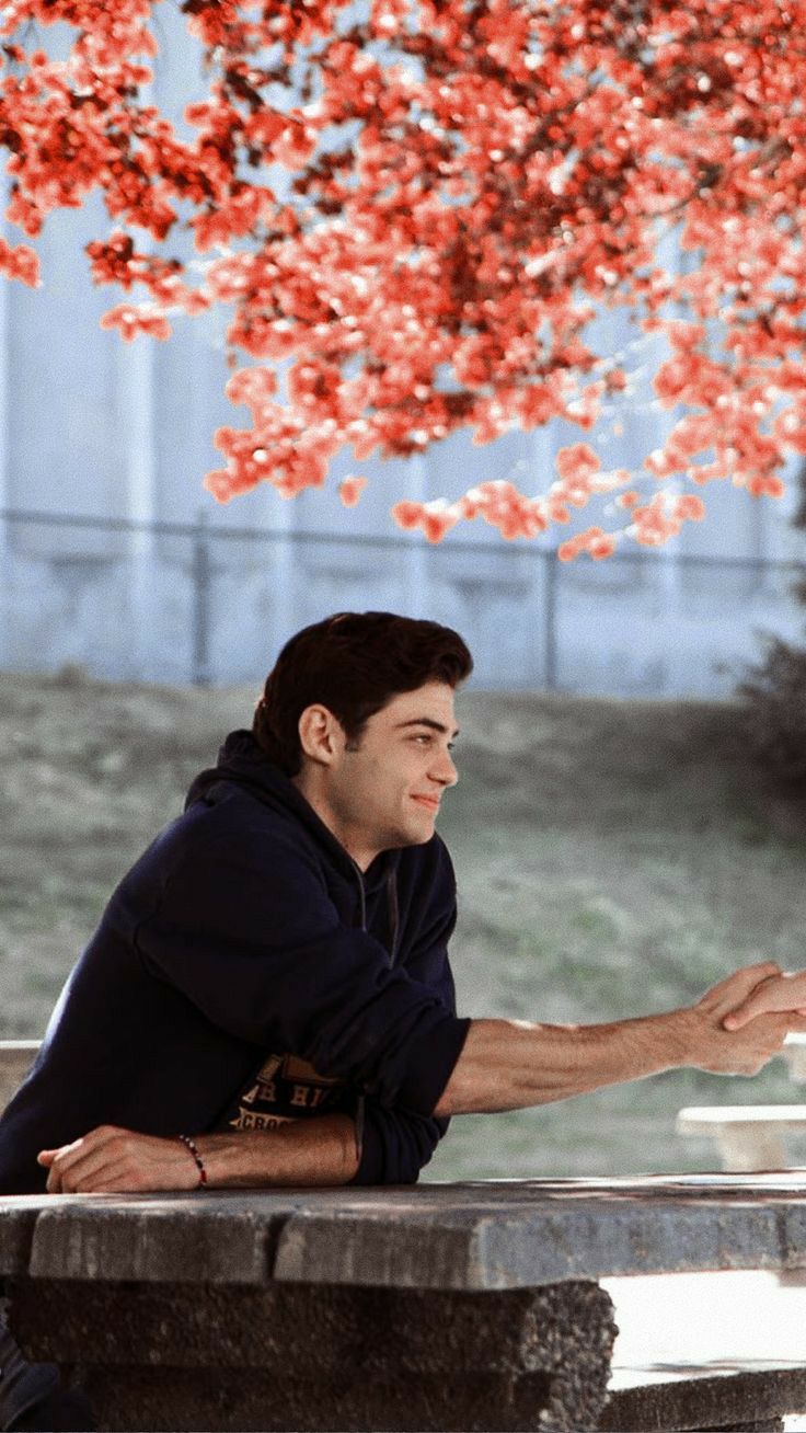 a man sitting on top of a cement bench under a tree with red leaves in the background