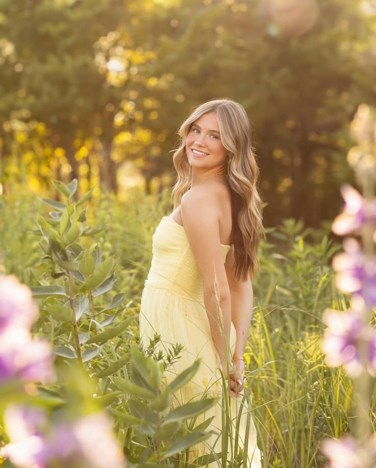 a beautiful woman in a yellow dress standing in tall grass with purple flowers behind her