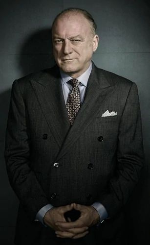 an older man in a suit and tie standing against a dark background with his hands together